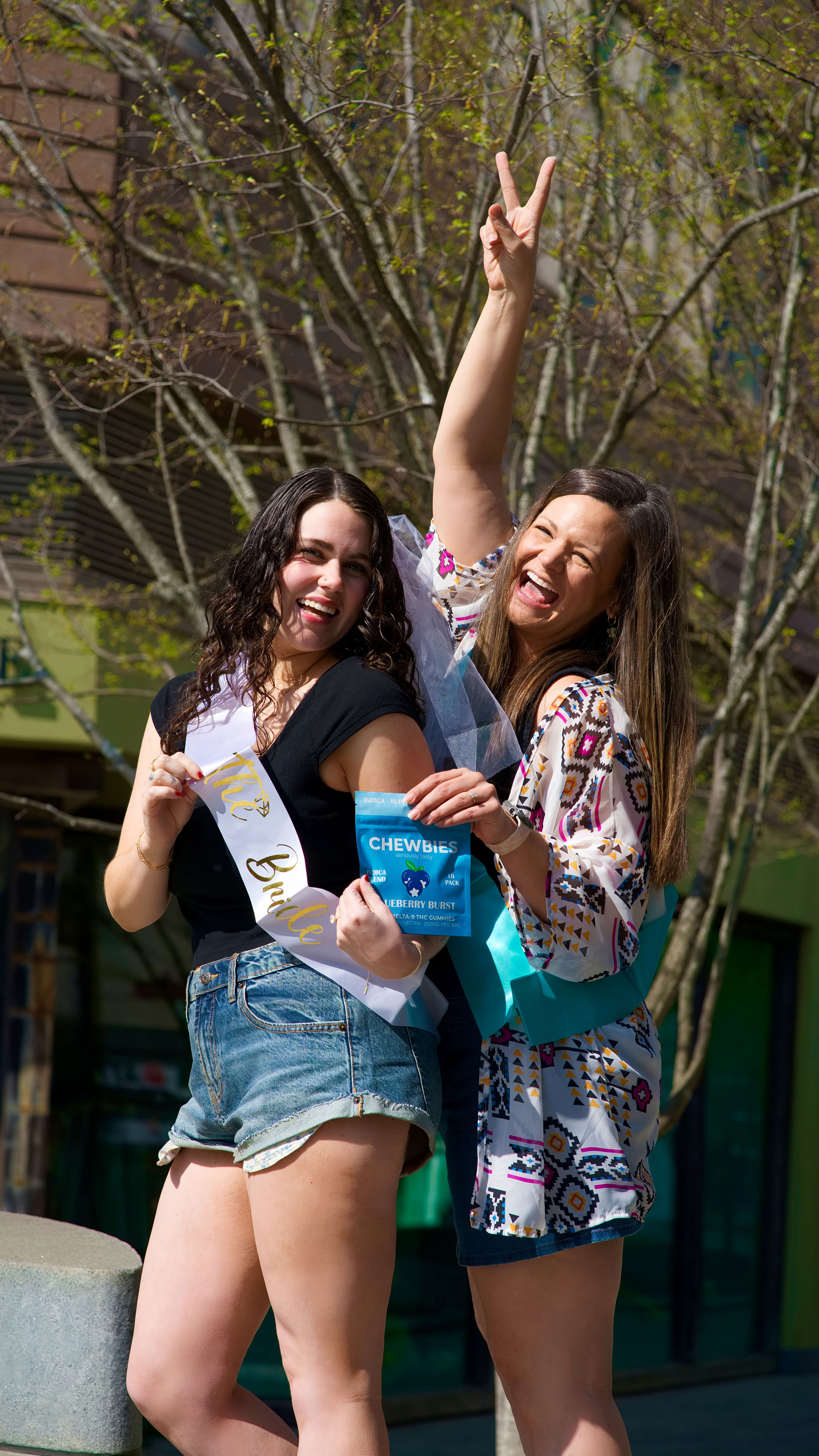 Two joyful women at a bachelorette celebration on a sunny spring downtown sidewalk — bride-to-be wearing a "Bride" sash and friend flashing a peace sign while holding a blue Chewbies pouch.
