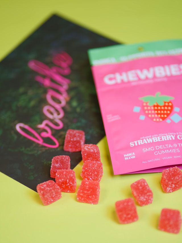 Vibrant close-up of sugar-coated strawberry gummy cubes spilling from a bright pink strawberry gummy pouch onto a yellow tabletop — colorful candy product shot.