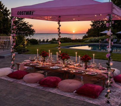 Romantic waterfront sunset dinner under a pink canopy with a low wooden table, velvet floor cushions, taper candles, red floral centerpieces and a poolside ocean view.