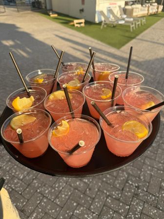 Sunlit tray of refreshing pink lemonade drinks in clear plastic cups with lemon wedges and black straws on an outdoor patio near poolside lounge chairs