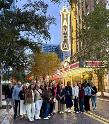 Smiling group of adults posing on a cobblestone downtown Tampa street at dusk beneath a glowing vintage theater marquee and neon sign, with trees and city buildings in the background.