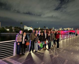Smiling group posing on a waterfront promenade at night with palm trees, city skyline and a red-lit bridge reflected in the river under a cloudy sky