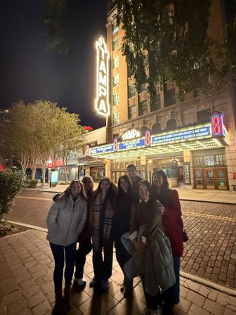 Group of friends smiling at night on a brick sidewalk in front of a lit historic Tampa theater marquee and vintage entrance