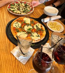 Thin-crust Margherita pizza with melted mozzarella and chopped basil on a black pan, alongside an arugula and shaved-parmesan carpaccio salad, lemon wedges, stacked plates and glasses of water and red wine drink on a wooden restaurant table