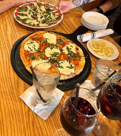 Thin-crust Margherita pizza with melted mozzarella and chopped basil on a black pan, alongside an arugula and shaved-parmesan carpaccio salad, lemon wedges, stacked plates and glasses of water and red wine drink on a wooden restaurant table