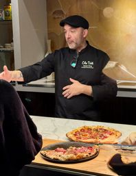 Hands-on chef in a black jacket and flat cap gesturing behind a marble counter in an open kitchen, two artisan Neapolitan-style pepperoni pizzas on a wooden peel under warm lighting — cozy pizzeria scene.