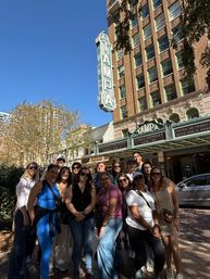Cheerful tour group posing beneath a vintage vertical "TAMPA" sign and ornate theater marquee in downtown Tampa on a sunny day