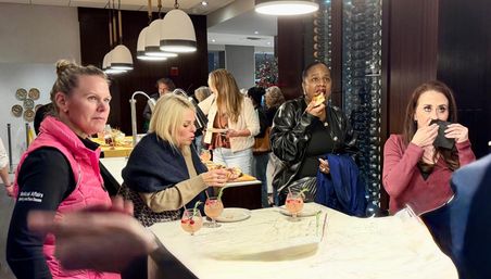 Women sampling appetizers and pink cocktails at an upscale hotel lounge bar with a marble countertop, pendant lights, wine wall and holiday decor.