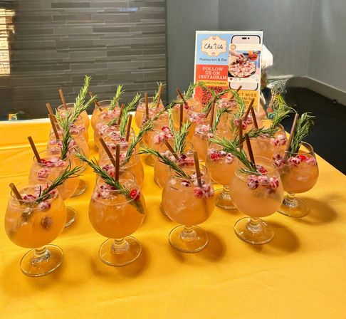 Peach-orange cocktails in stemmed glasses arranged on a yellow tablecloth, each garnished with rosemary sprigs, sugared cranberries and cinnamon sticks for a festive restaurant event.