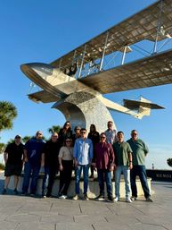 Group of people posing in front of a large historic seaplane monument mounted on a curved pedestal at a sunny waterfront plaza with palm trees and clear blue sky