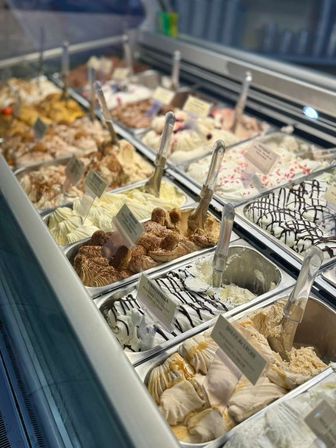 Mouthwatering artisan gelato display case with multiple flavored pans — chocolate-drizzled, caramel-swirled and sprinkled varieties with clear scoops in a glass counter.