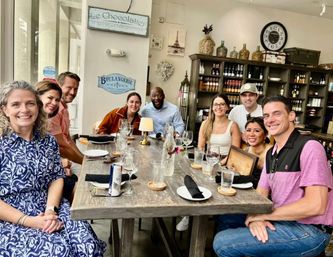 Smiling group of friends seated around a rustic wooden table in a French‑style bistro with wine shelves, glasses and plates ready for brunch.