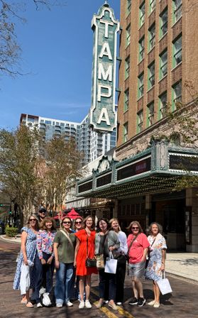 Cheerful group of adults posing on a sunny downtown Tampa street beneath a tall vintage green theater marquee and ornate brick facade.