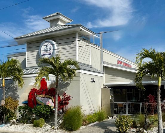 Tropical seafood restaurant exterior with white coastal-style building and metal roof, palm trees flanking an outdoor patio, and a large bright red lobster mural on the wall under a blue sky.
