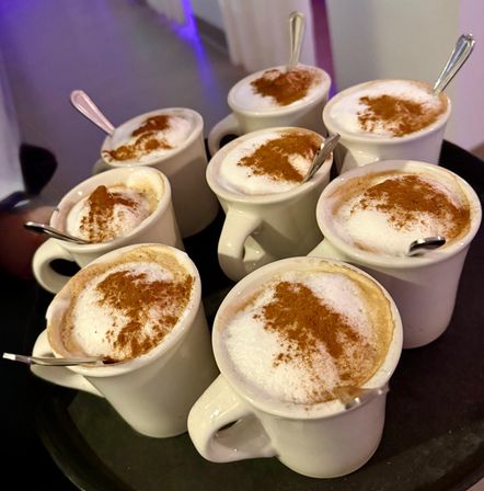 Eight frothy cappuccinos in white ceramic mugs, each topped with frothed milk and a cinnamon dusting, arranged on a black serving tray.