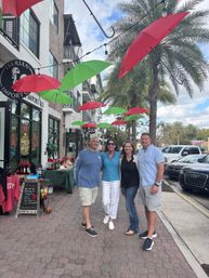 Four adults smiling for a photo under hanging red and green umbrellas on a palm-lined downtown shopping street with storefront displays and festive holiday decor.