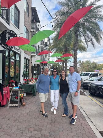 Four adults smiling for a photo under hanging red and green umbrellas on a palm-lined downtown shopping street with storefront displays and festive holiday decor.