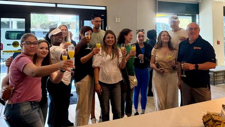 Group of diverse adults smiling and toasting with mimosas inside a bright modern cafe or hotel lobby during a daytime brunch