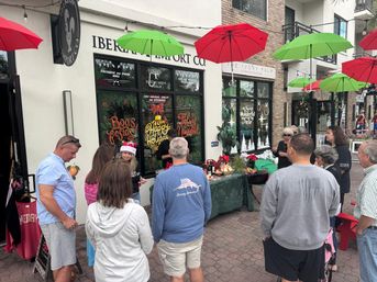 Outdoor holiday market scene with shoppers gathered around a festive shopfront and vendor table on a pedestrian shopping street, red and green umbrellas suspended overhead, holiday window signs and poinsettias adding seasonal decor.