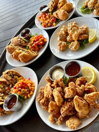 Assorted appetizer plates: crispy fried chicken and seafood bites, crunchy egg-roll style wraps, bright mango salsa, dipping sauces and lemon wedges on white plates on a restaurant patio tray.