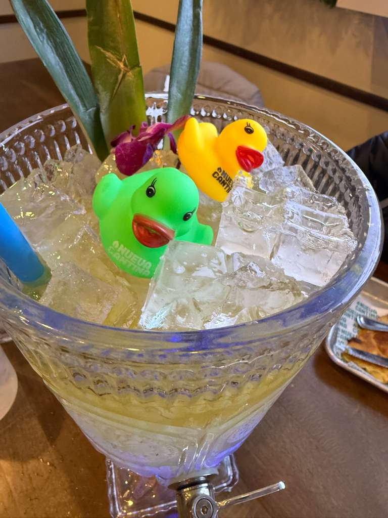 Playful ice-filled glass beverage dispenser with green and yellow rubber ducks, a purple orchid and tropical leaves used as garnish on a wooden table.