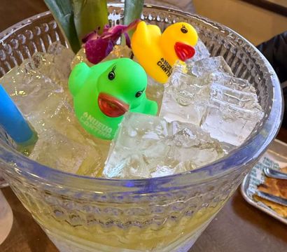 Playful ice-filled glass beverage dispenser with green and yellow rubber ducks, a purple orchid and tropical leaves used as garnish on a wooden table.