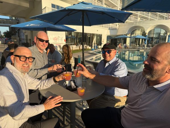Four men toasting fruity cocktails at a sunny poolside patio with blue umbrellas, lounge chairs and a hotel-style building in the background — casual outdoor bar vibe.