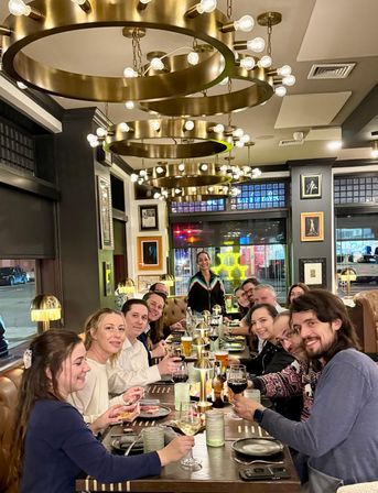 Group dinner in a cozy urban restaurant at night — a long communal table of smiling people toasting with wine and beer beneath large circular brass chandeliers and warm table lamps, city street visible through windows.