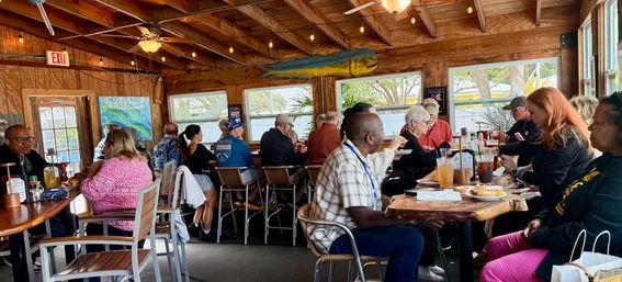 Sunlit beachside restaurant with rustic wood interior and a large painted fish, busy bar and tables of patrons enjoying brunch plates and drinks.