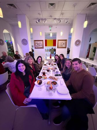 Eight adults smiling around a long white table in a modern, brightly lit restaurant with pendant lights, tapas plates, bread and sangria glasses for a lively group dinner