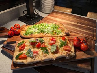 Two rectangular flatbread pizzas with charred crusts on wooden serving boards, topped with fresh arugula, basil, cherry tomato halves and grated cheese, with vine tomatoes on the side on a kitchen countertop.