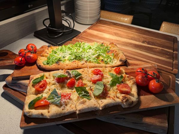 Two rectangular flatbread pizzas with charred crusts on wooden serving boards, topped with fresh arugula, basil, cherry tomato halves and grated cheese, with vine tomatoes on the side on a kitchen countertop.