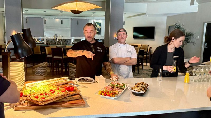 Hotel lounge buffet counter with staff presenting a brunch spread: flatbreads topped with cherry tomatoes, colorful salad skewers, grilled bread in a bowl, stacked plates, carafes of orange juice and wine glasses under warm pendant lighting.