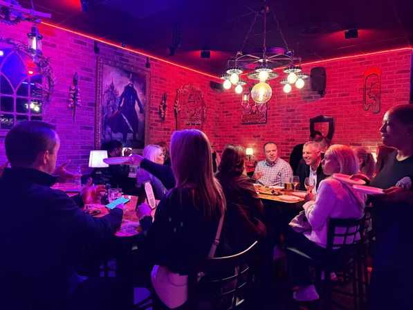 Lively evening inside a cozy brick-walled pub: a group gathered at a long table under red neon lights and a vintage chandelier, sharing plates, drinks and conversation.