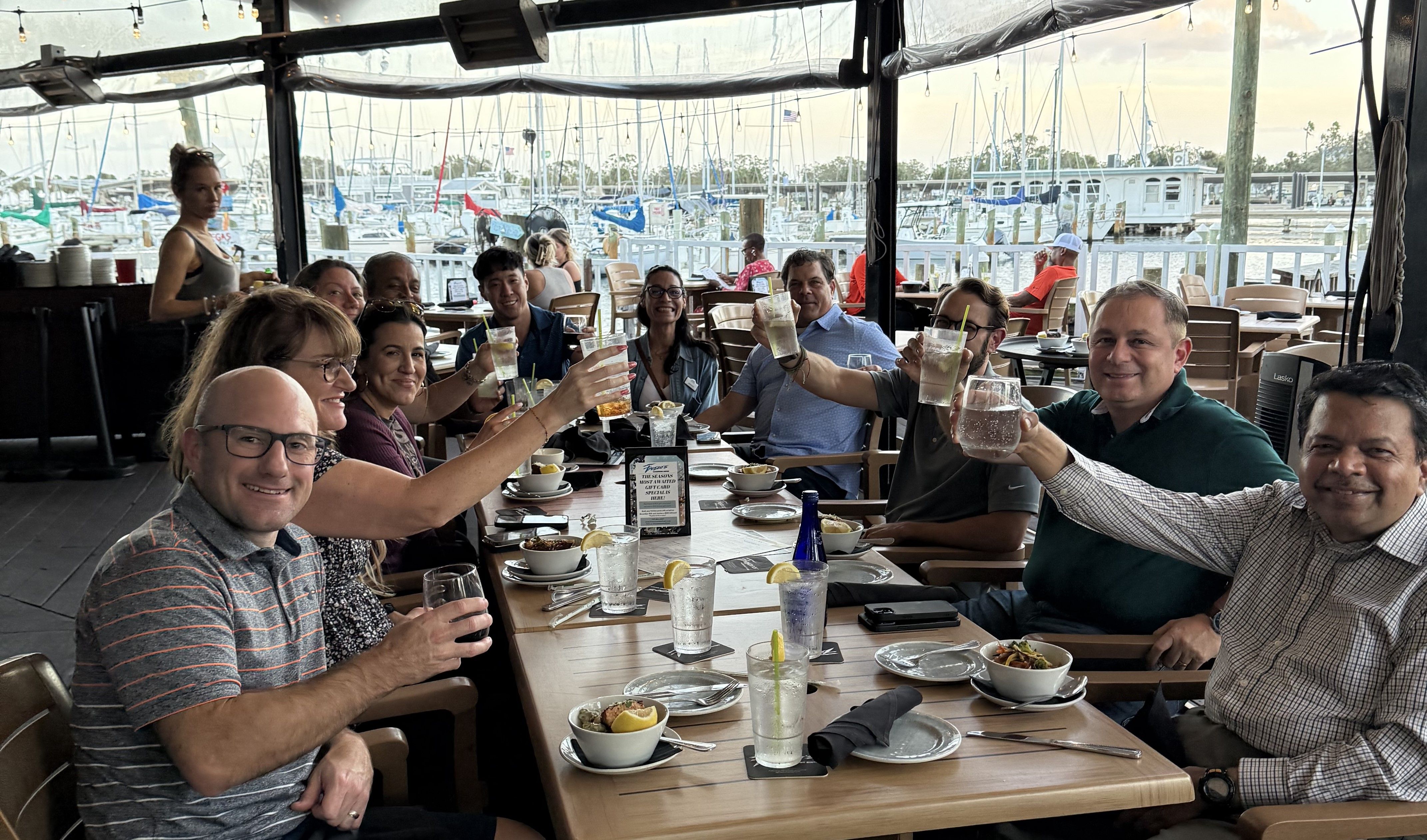 Cheerful group of adults toasting with drinks at a waterfront restaurant patio, wooden table set with bowls and glasses, overlooking a marina filled with sailboats and docks.