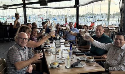 Cheerful group of adults toasting with drinks at a waterfront restaurant patio, wooden table set with bowls and glasses, overlooking a marina filled with sailboats and docks.