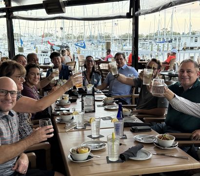 Cheerful group of adults toasting with drinks at a waterfront restaurant patio, wooden table set with bowls and glasses, overlooking a marina filled with sailboats and docks.