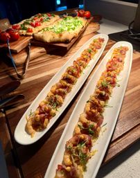 Two long white platters of bite-sized bacon-wrapped shrimp appetizers with microgreens and mustard dots on a wooden serving board, flatbread topped with greens and cherry tomatoes visible in the background