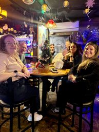 Group of friends smiling around a high-top table with a small Christmas tree reading "LOVE" in a cozy, colorful holiday-themed neighborhood bar with hanging lamps and a disco ball.