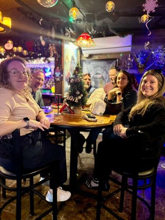 Group of friends smiling around a high-top table with a small Christmas tree reading "LOVE" in a cozy, colorful holiday-themed neighborhood bar with hanging lamps and a disco ball.