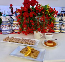 Spanish dessert spread with sugar-dusted churro sticks, caramel flan, hot chocolate and coffee, plus grilled crepes with orange slices, set against vibrant red flowers and hand-painted Spanish-style ceramic pitchers.