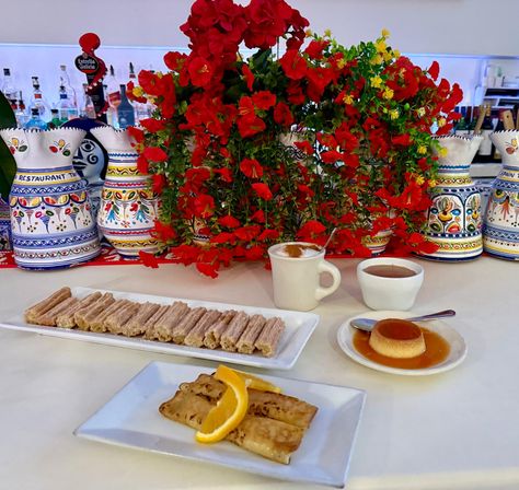 Spanish dessert spread with sugar-dusted churro sticks, caramel flan, hot chocolate and coffee, plus grilled crepes with orange slices, set against vibrant red flowers and hand-painted Spanish-style ceramic pitchers.