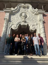 Smiling group of adults posing on stone steps beneath an ornate white carved Baroque-style archway set in a pale pink stucco facade on a sunny day