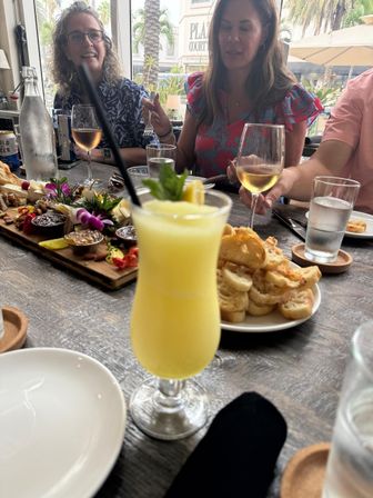 Frozen yellow tropical cocktail with mint and lemon in foreground at a coastal patio brunch table with charcuterie board, crostini, wine glasses and people chatting, palm trees visible outside.