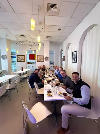 Smiling group enjoying a communal dinner at a long white table in a bright modern restaurant with hanging pendant lights, arched alcoves, minimalist white chairs and small plates of food.