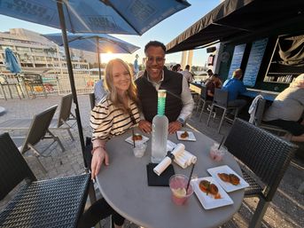 Two people smiling at a waterfront outdoor patio bar table with cocktails, small plates and a tall bottle under blue umbrellas at sunset