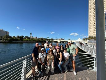 Smiling group posing on a sunny downtown riverwalk promenade with palm trees, a pedestrian bridge, high-rise buildings and calm waterfront