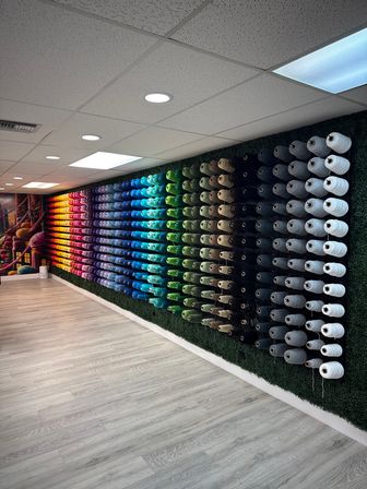 Vibrant indoor craft store wall of hundreds of yarn spools arranged in a rainbow gradient from warm reds through blues to neutral whites on a green backdrop, lit by recessed ceiling lights over light wood flooring.