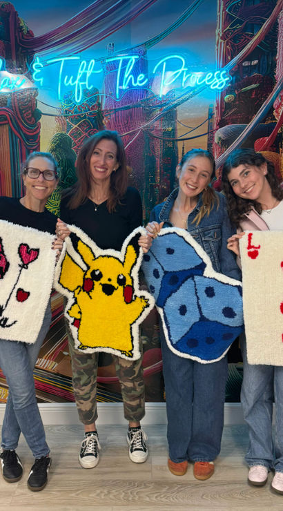 Four participants in a colorful tufting workshop holding handmade tufted rugs — pink poodle with hearts, yellow Pikachu, blue dice, and a red-hearts playing card against a neon-lit mural backdrop.