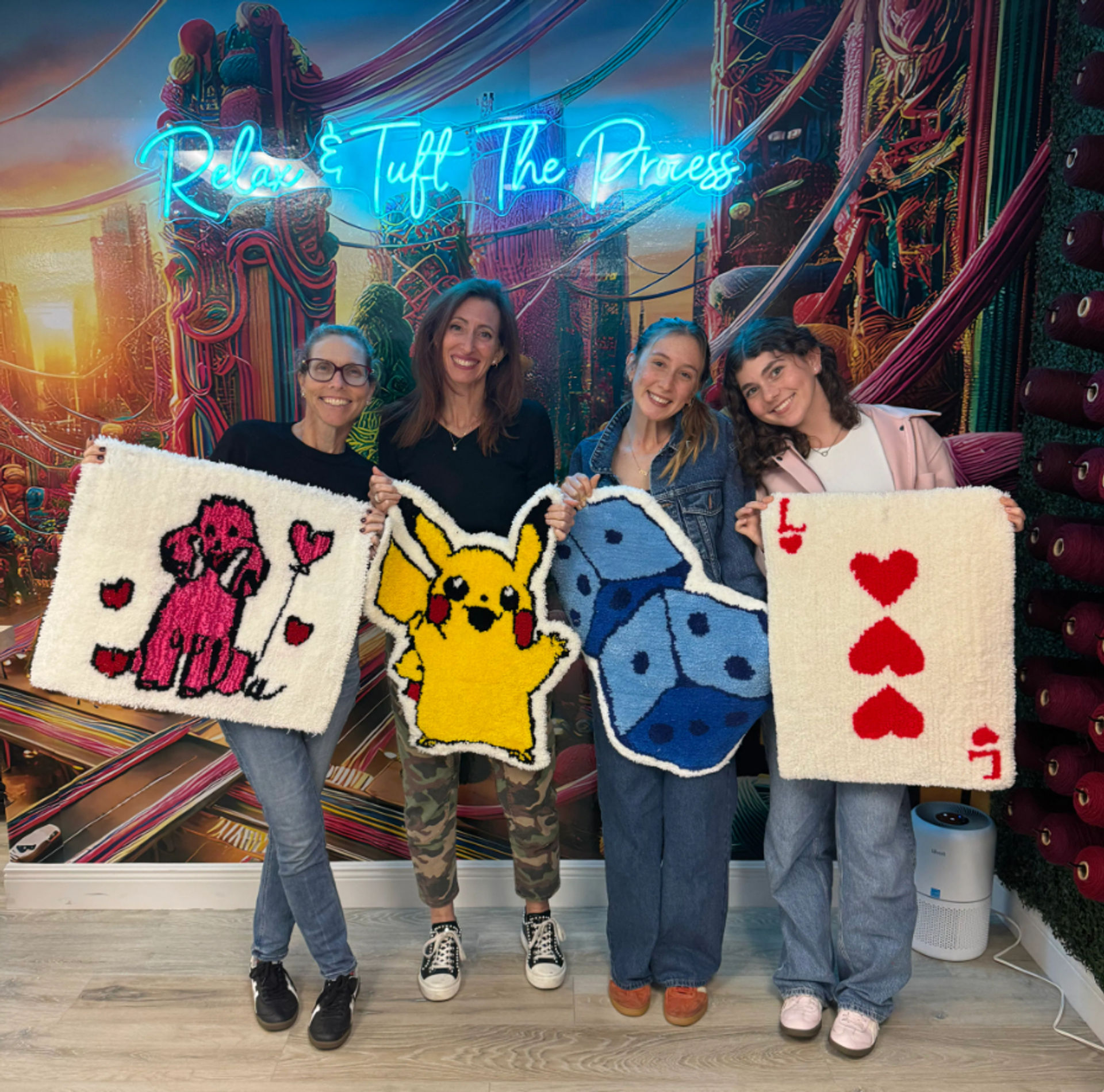 Four participants in a colorful tufting workshop holding handmade tufted rugs — pink poodle with hearts, yellow Pikachu, blue dice, and a red-hearts playing card against a neon-lit mural backdrop.